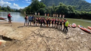 Un grupo de personas de pie en una playa arenosa sosteniendo remos, con un río y montañas al fondo.