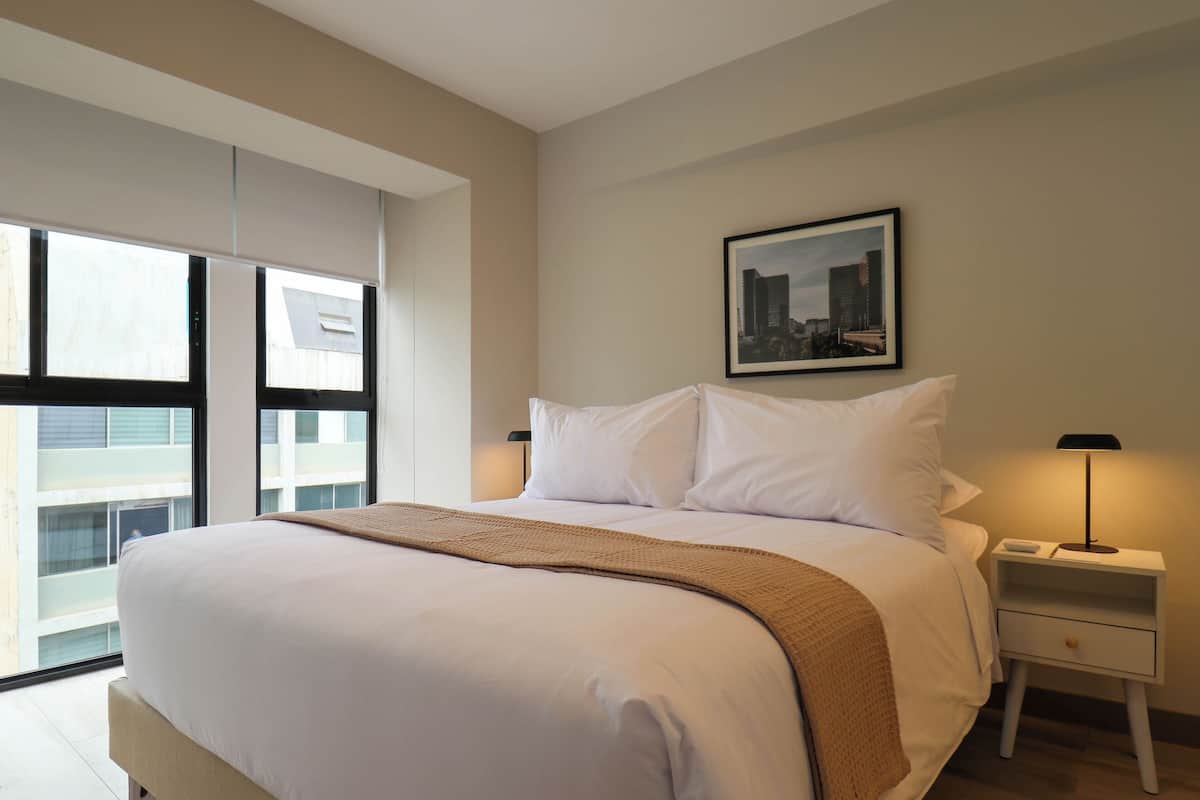 Quarto de hotel moderno com uma cama grande, uma mesa de cabeceira com abajur e vista para a cidade através de amplas janelas.