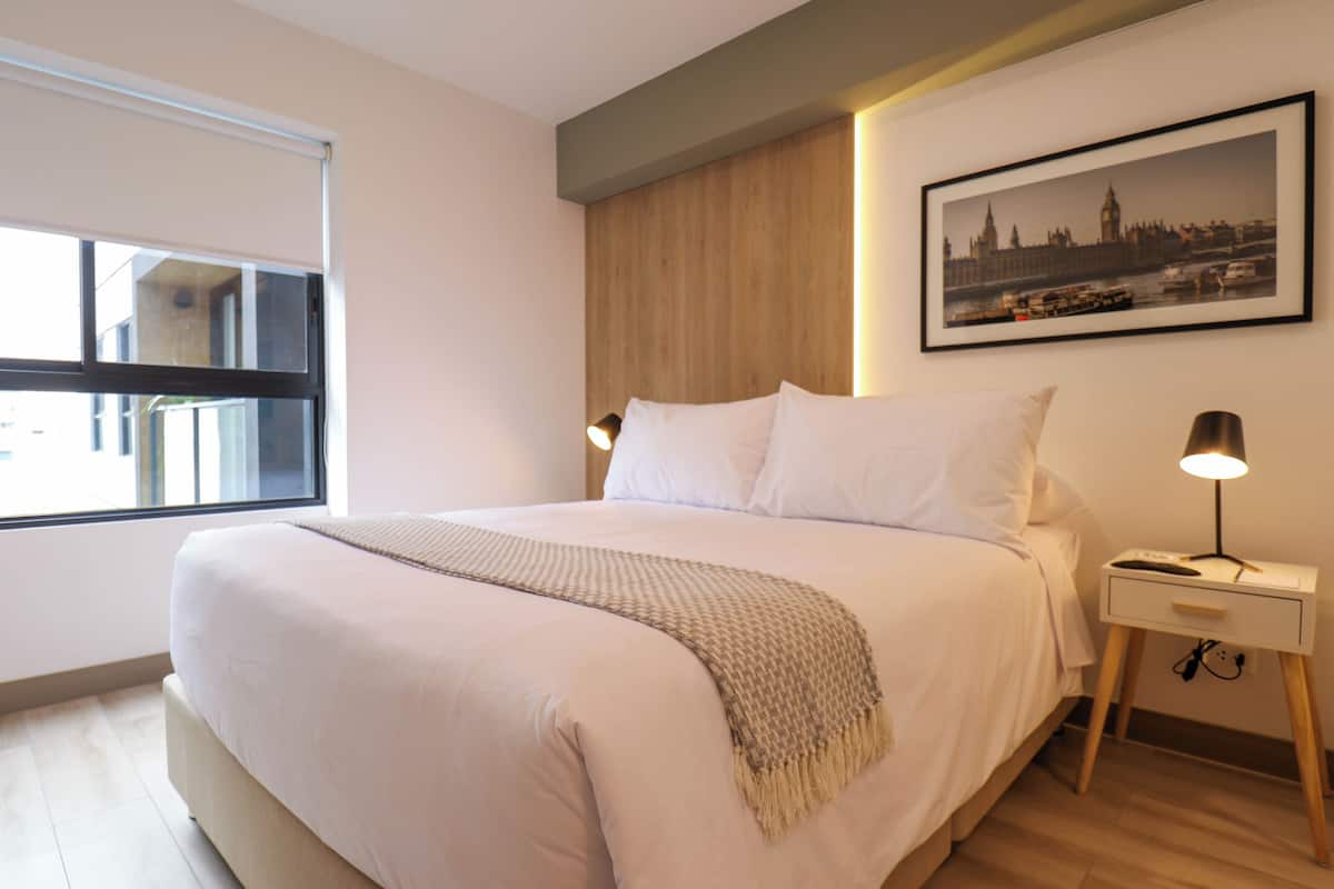 Uma cama bem arrumada com um edredom branco e um cobertor bege, uma mesa de cabeceira com abajur e um quadro com uma vista da cidade na parede.