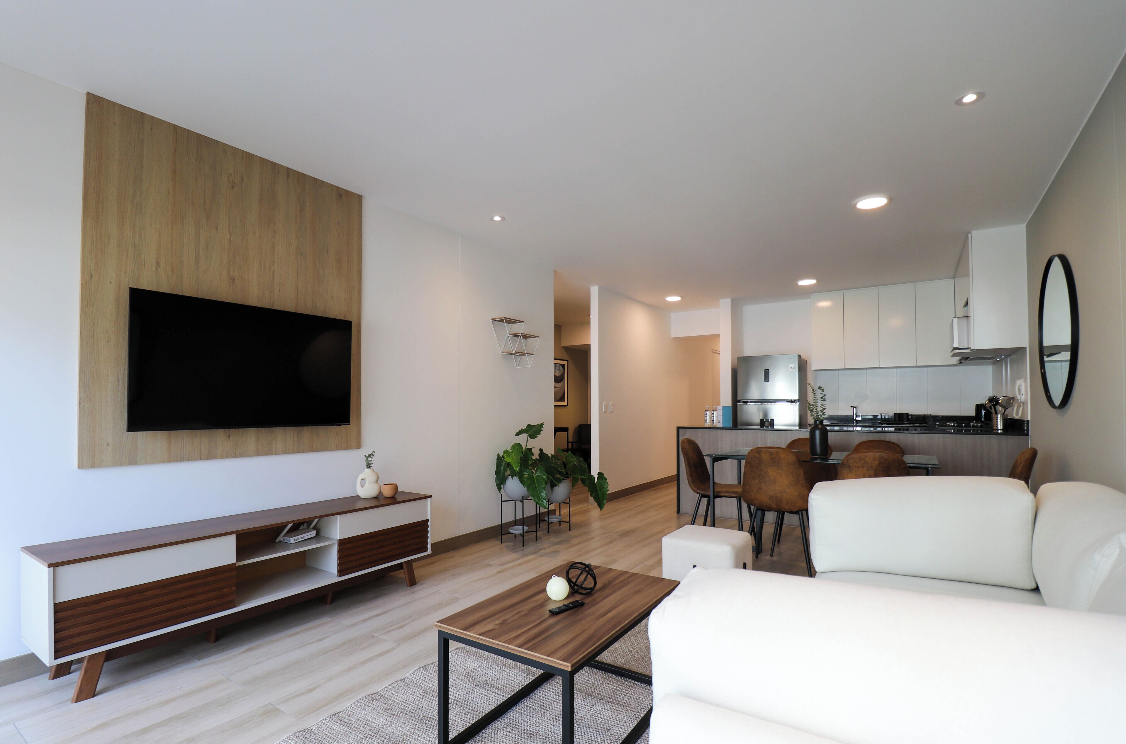 Una sala de estar moderna con una televisión de pantalla plana, un sofá, una mesa de centro y un comedor con sillas, y una cocina al fondo.