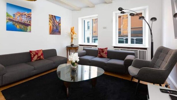 En moderne stue med en grå sofa, et rundt sofabord og en sort måtte. To billeder hænger på væggen, der er en gulvlampe, og et vindue med udsigt til bygninger.