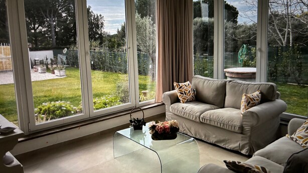 En moderne stue med et glasbord, en beige sofa og udsigt til en have gennem store vinduer.
