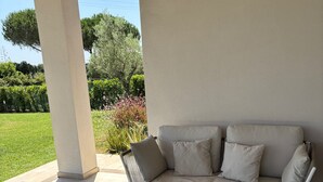 En overdækket terrasse med en hvid sofa, et rundt bord og en træskål.