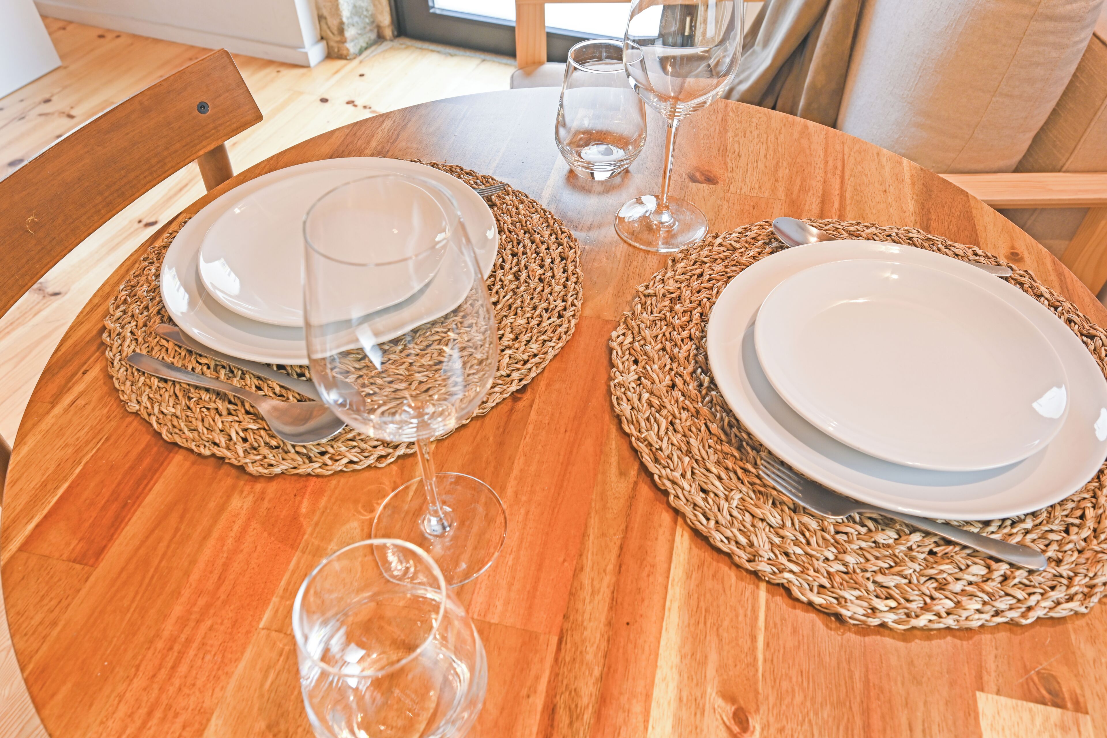 Una mesa redonda de madera con individuales tejidos, platos blancos y copas de vino dispuestas para comer.