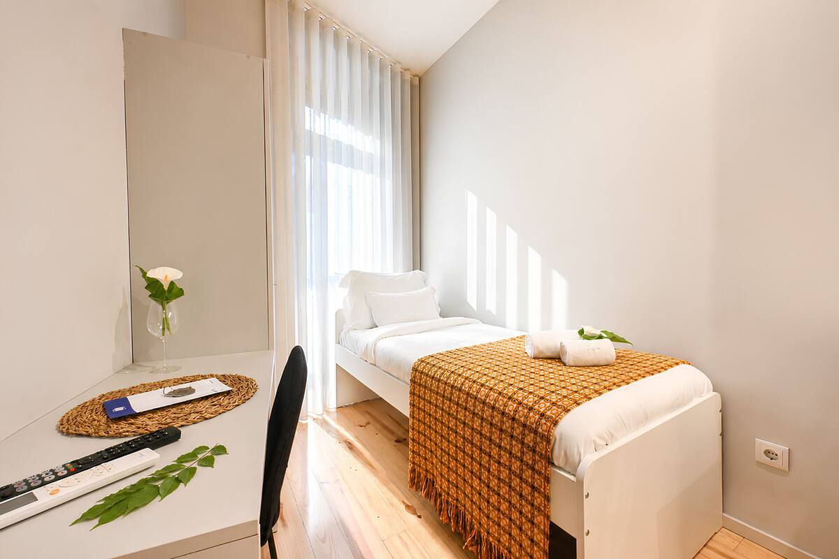 A small, neatly arranged bedroom with a bed, a desk with a computer, a chair, and a window with curtains.