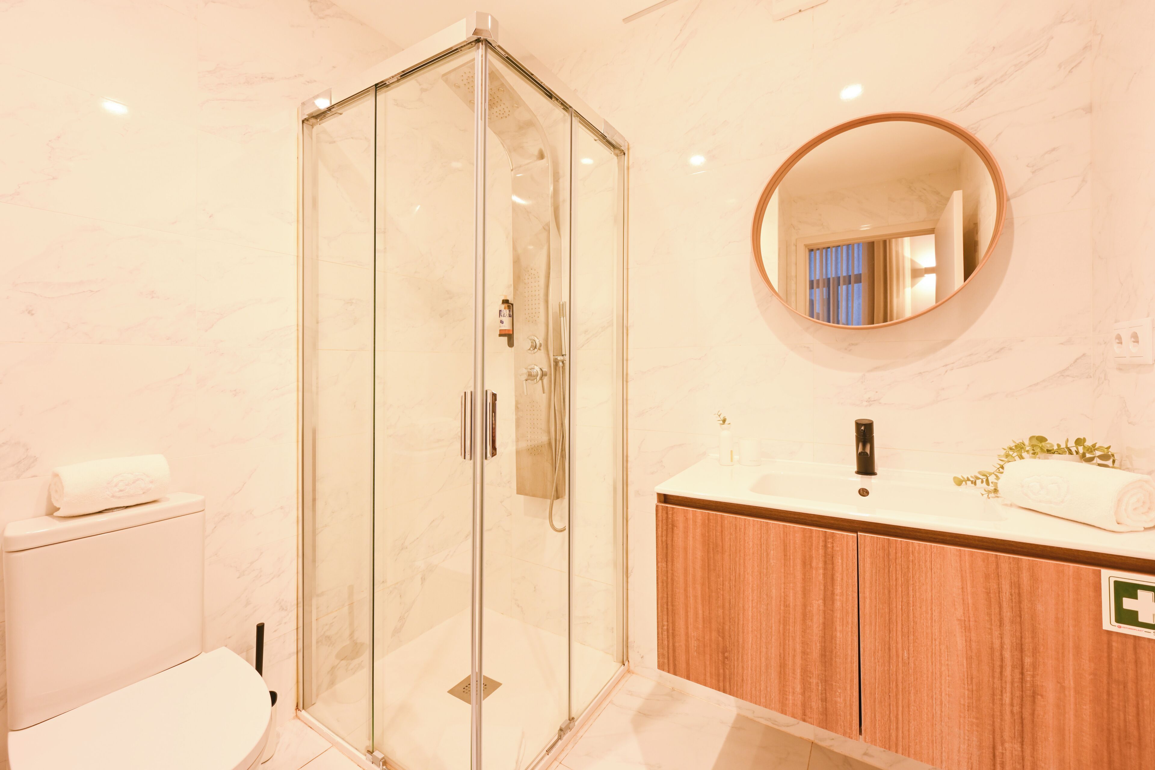 Un baño moderno con cabina de ducha de vidrio, tocador de madera, espejo redondo y inodoro a la vista.