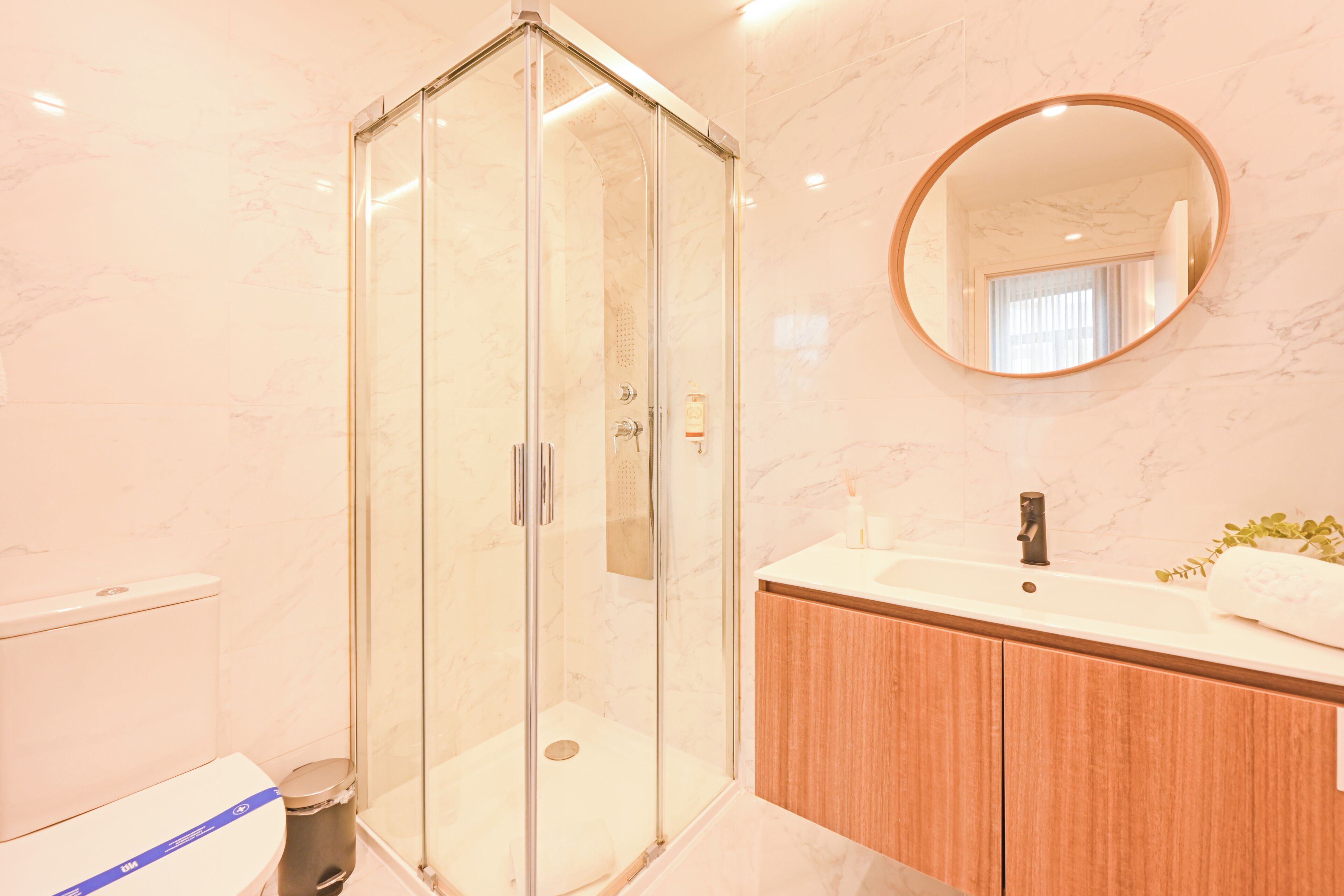 A modern bathroom with a glass-enclosed shower, a wooden vanity, and a round mirror.