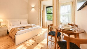 A bedroom with a bed, bedside table, lamp, and a dining area with a table, chairs, and a window. - FindMe inPorto Bonfim (Porto)