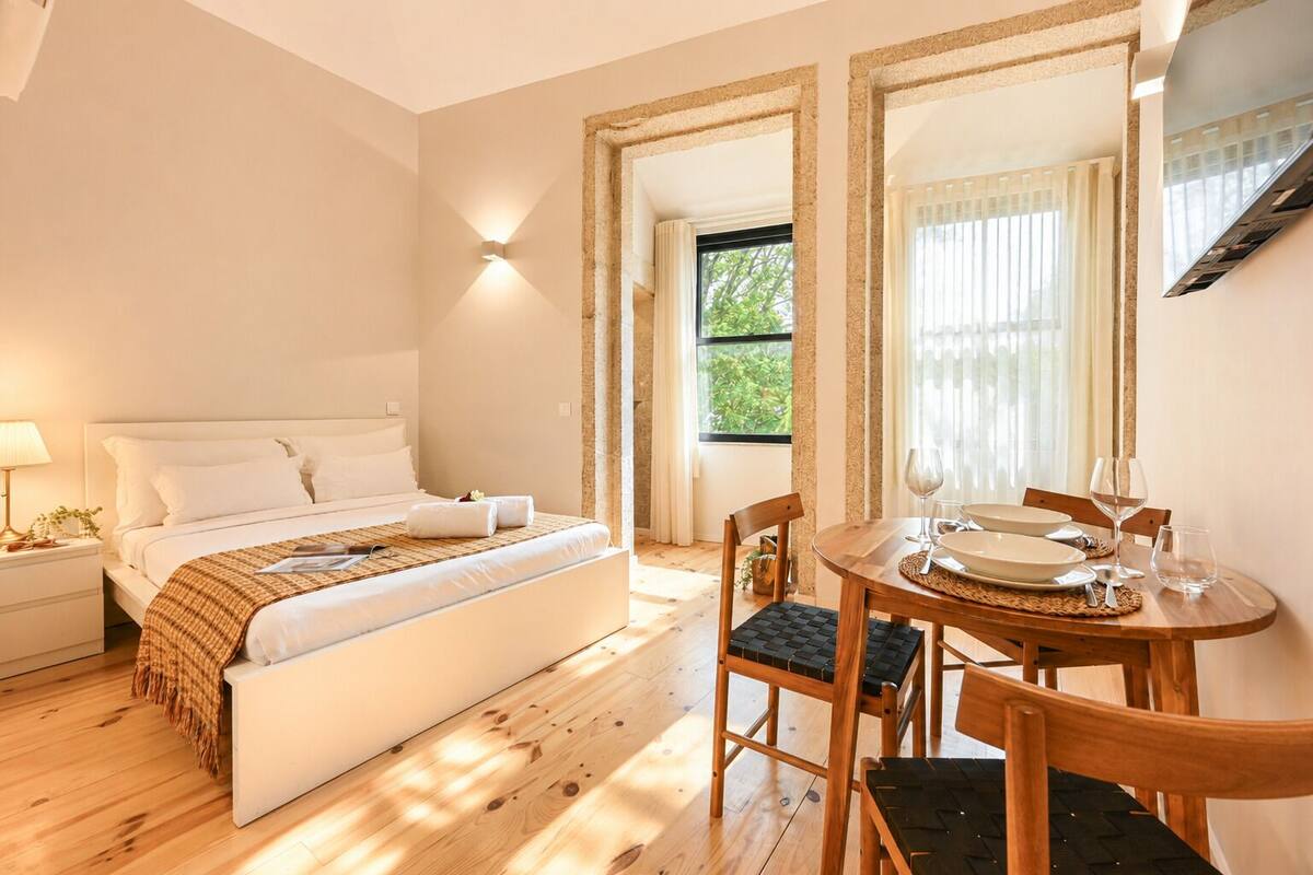 A bedroom with a bed, bedside table, lamp, and a dining area with a table, chairs, and a window.