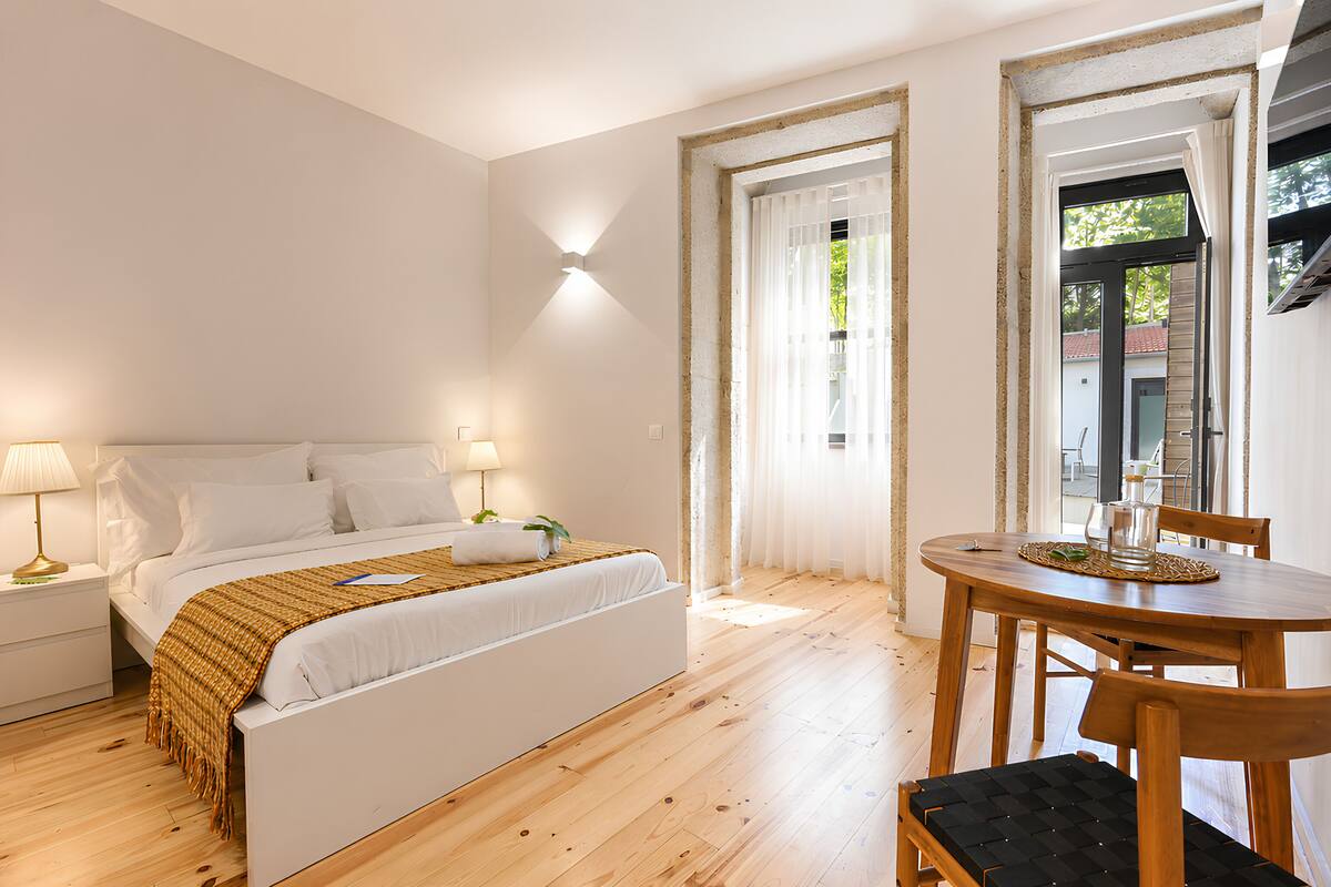 A bedroom with a bed, bedside tables, a wooden floor, a window, and a dining table with chairs.