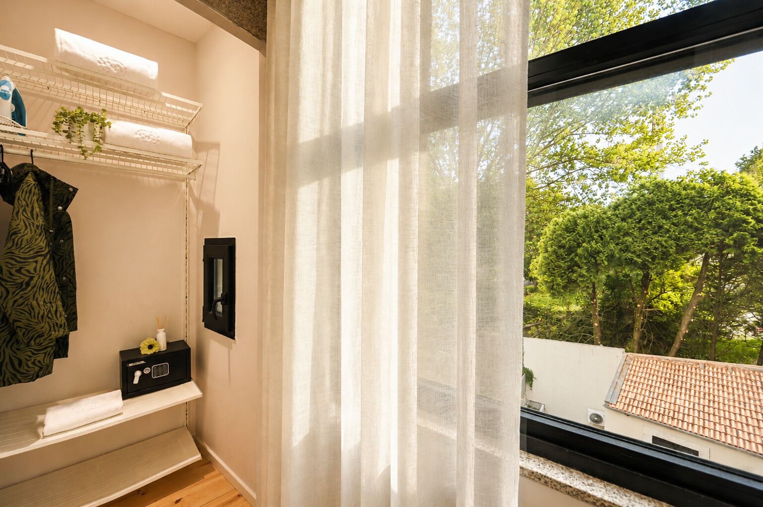 A room with a window, a radiator, a small table with a plant, and a shelf with towels and a coat.