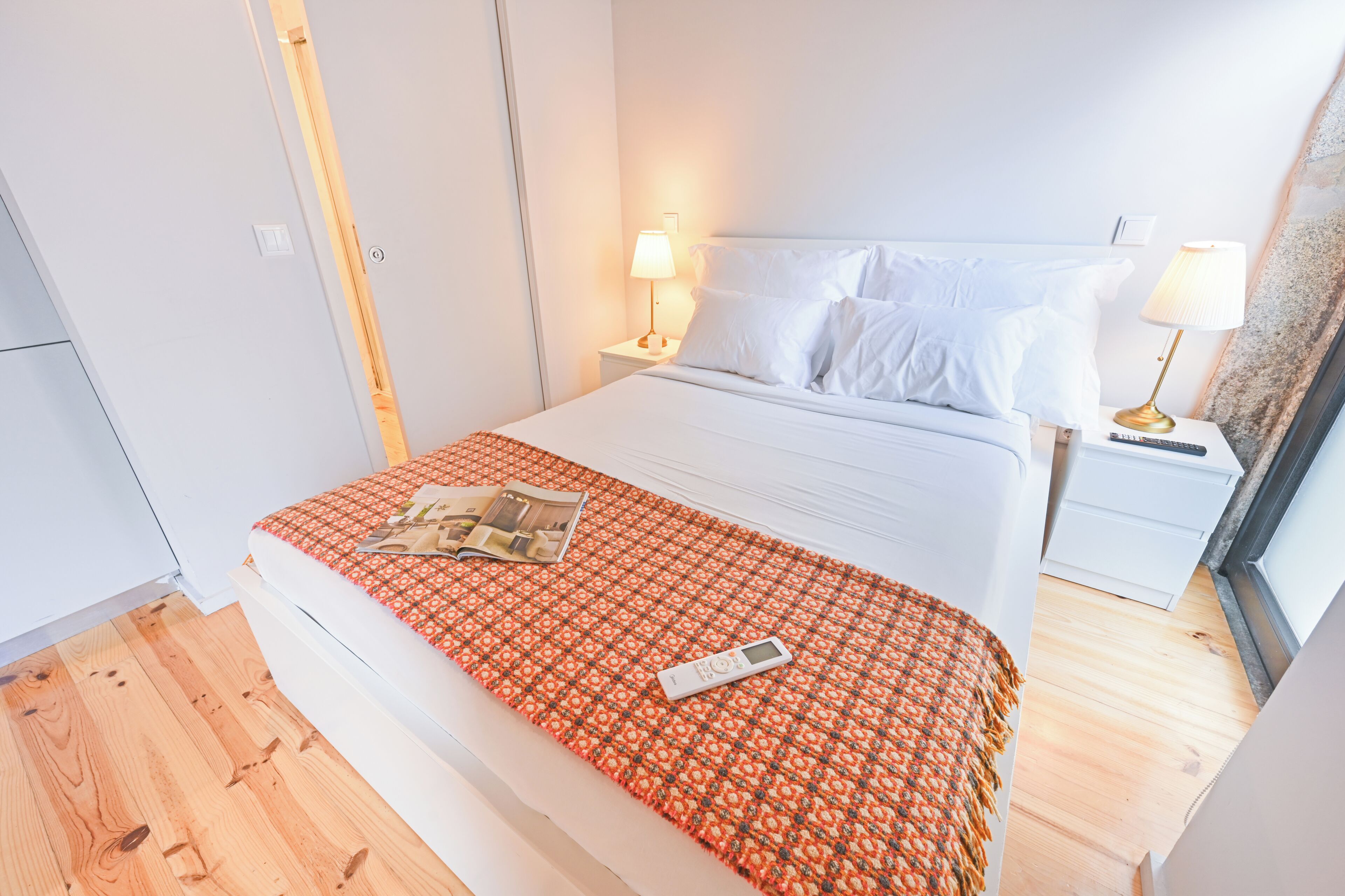 A hotel room with a bed, a nightstand with a lamp, a remote control, and a magazine on the bed.