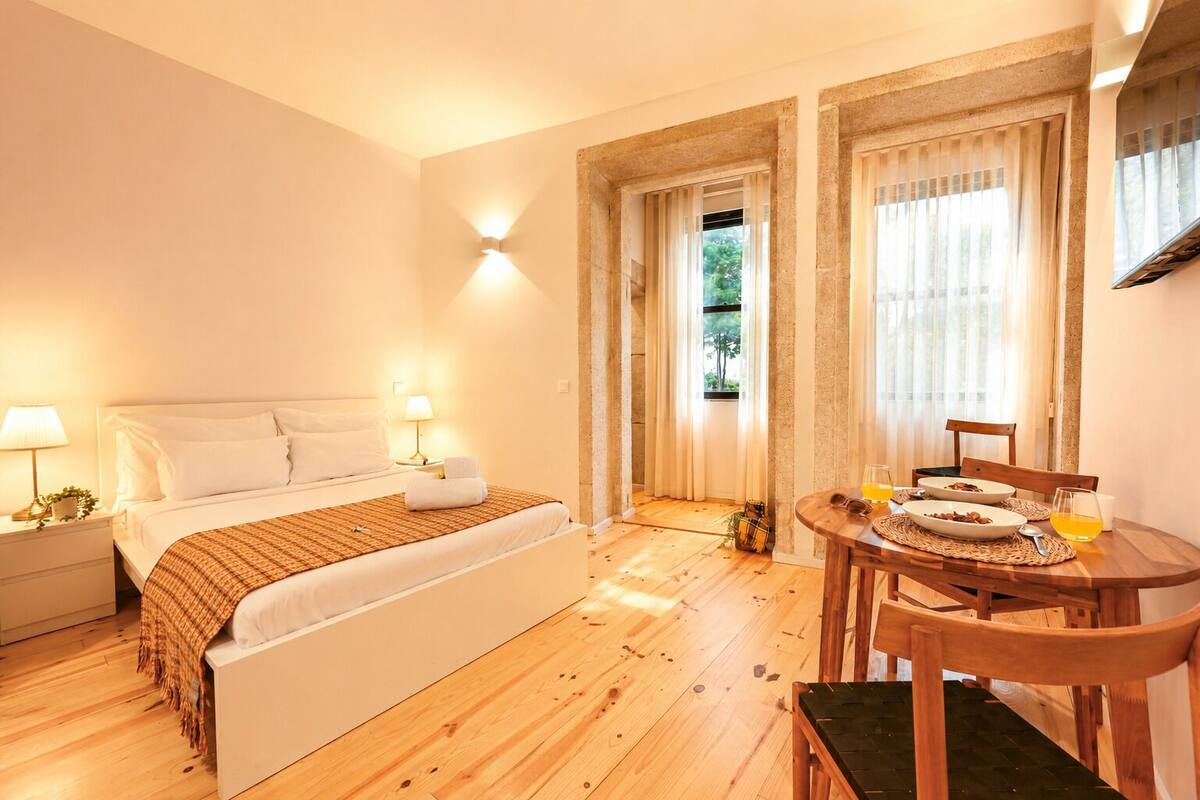 A bedroom with a bed, bedside lamps, a wooden floor, a dining table with chairs, and a window with curtains.