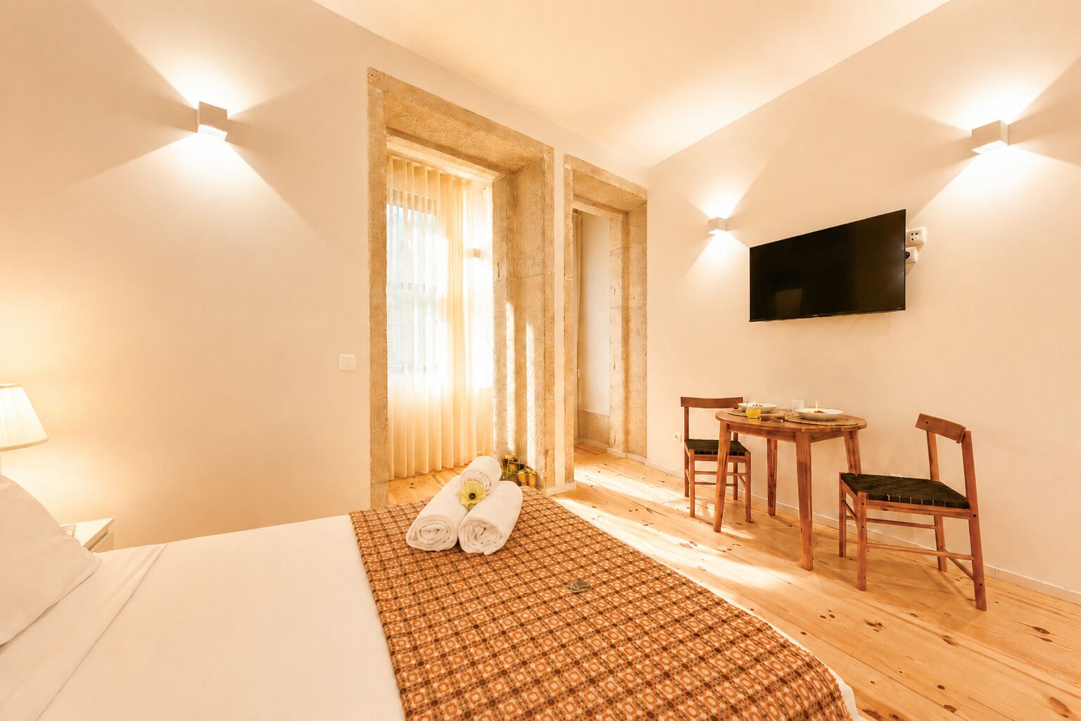 A hotel room with a bed, a desk, two chairs, a TV, and a window with curtains.