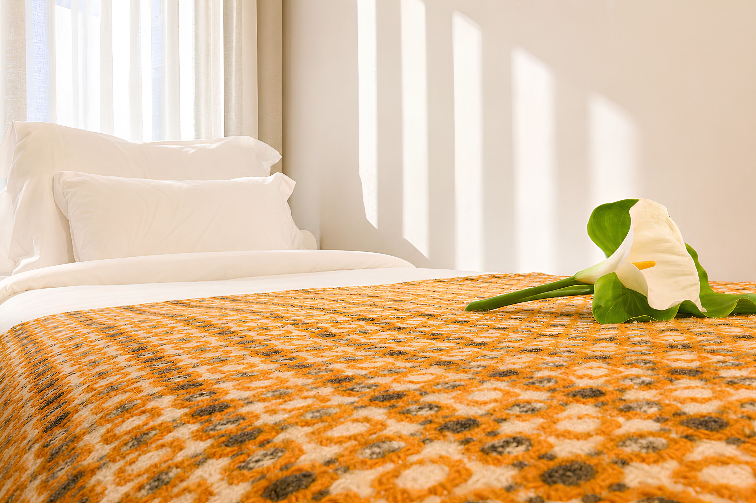 A neatly made bed with white linens, a patterned orange and gray blanket, and a single white flower on the bed.