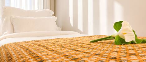 A neatly made bed with white linens, a patterned orange and gray blanket, and a single white flower on the bed.