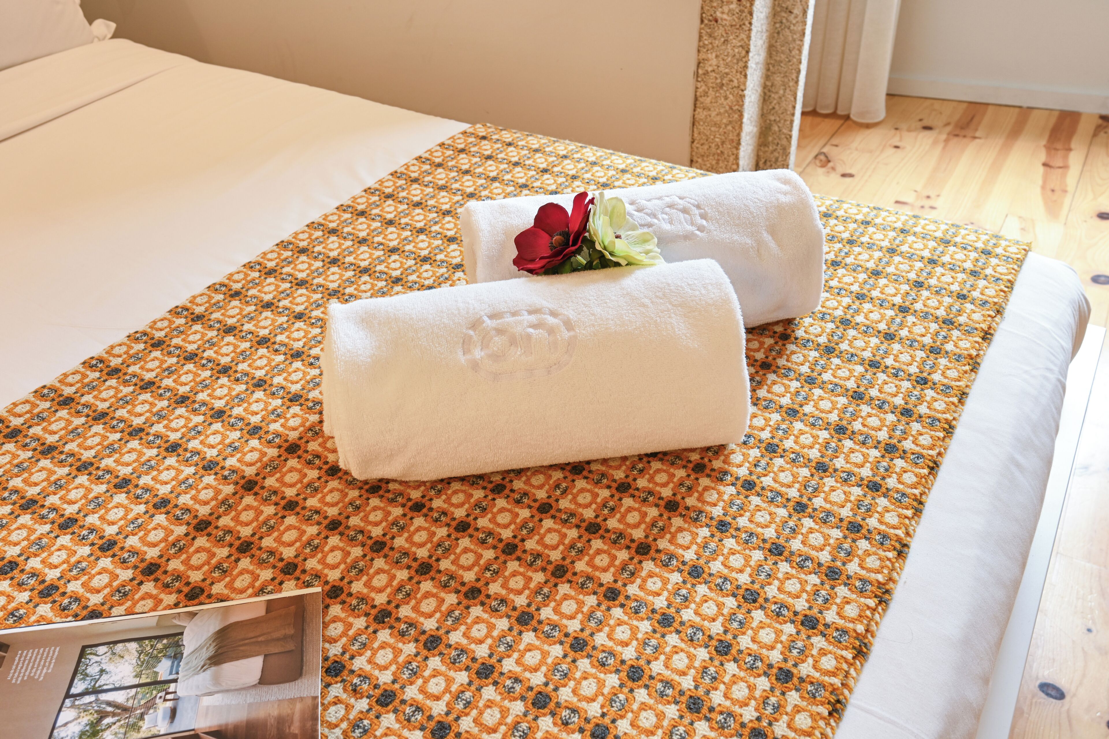 Una cama bien hecha con una colcha estampada, dos toallas enrolladas con una flor roja y un cuadro en el lateral.