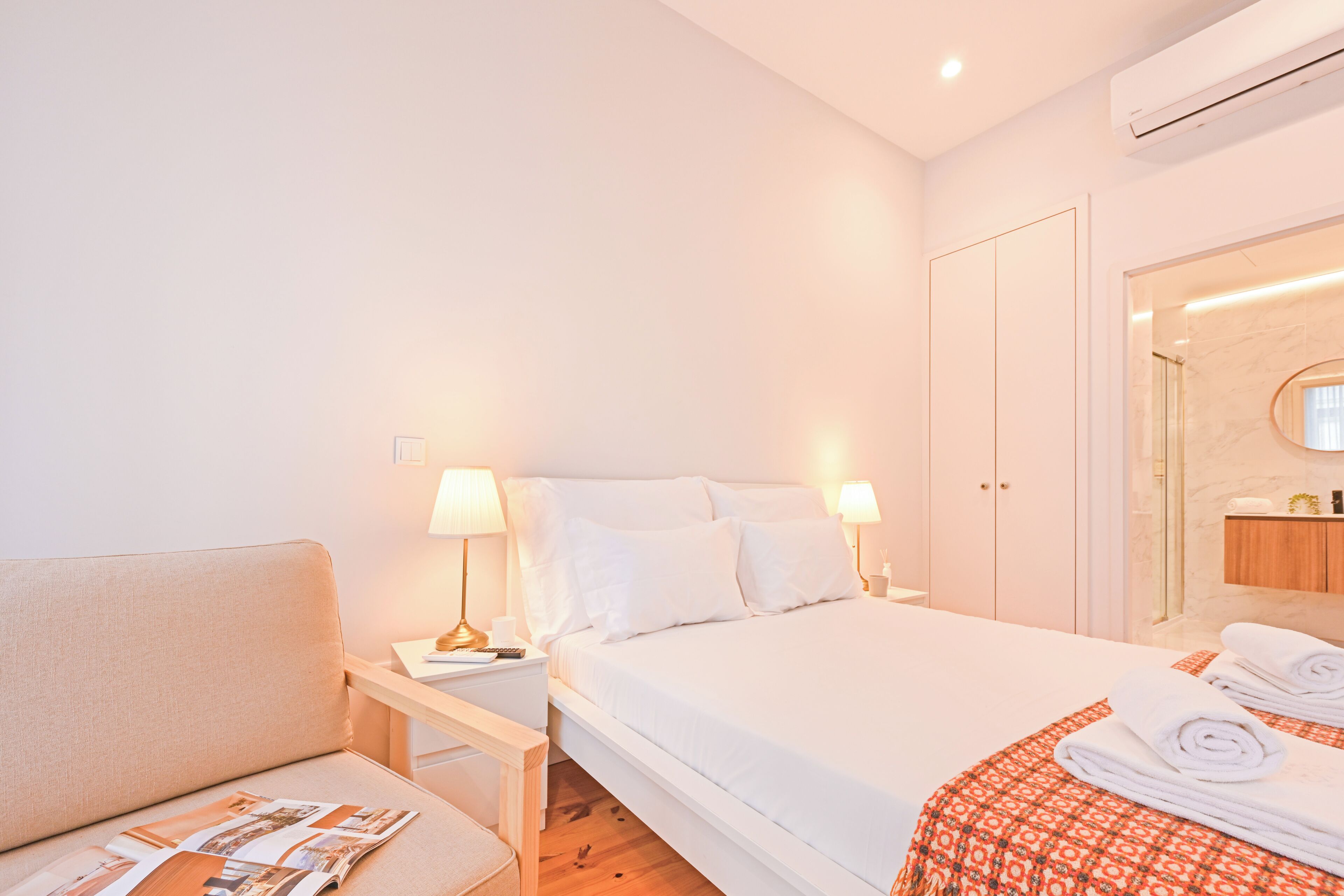 A hotel room with a bed, bedside tables, a chair, a mirror, and a bathroom visible through an open door.