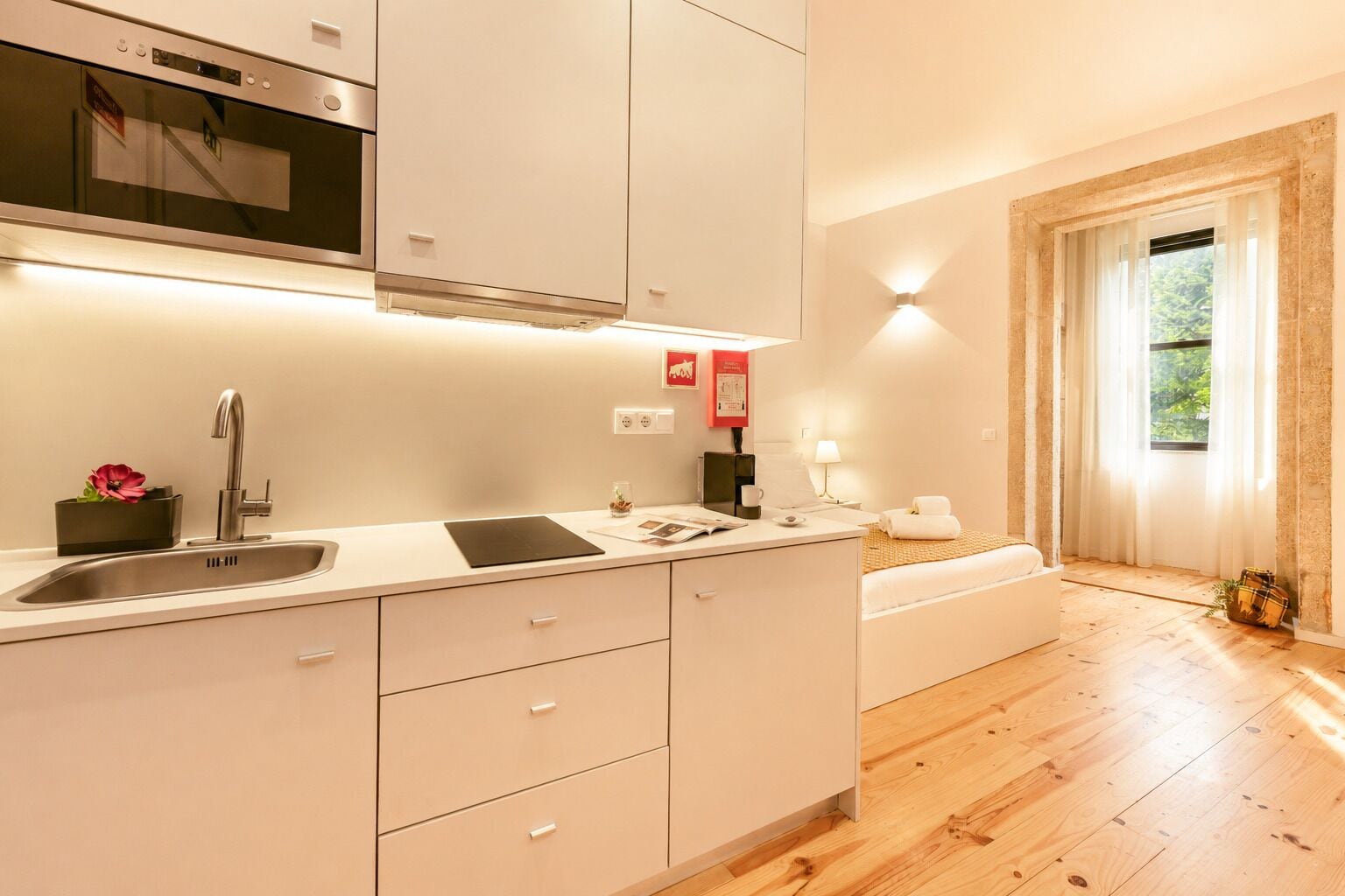 A modern kitchen with a built-in oven, a sink, and a countertop. There is a window with curtains and a view of greenery outside.
