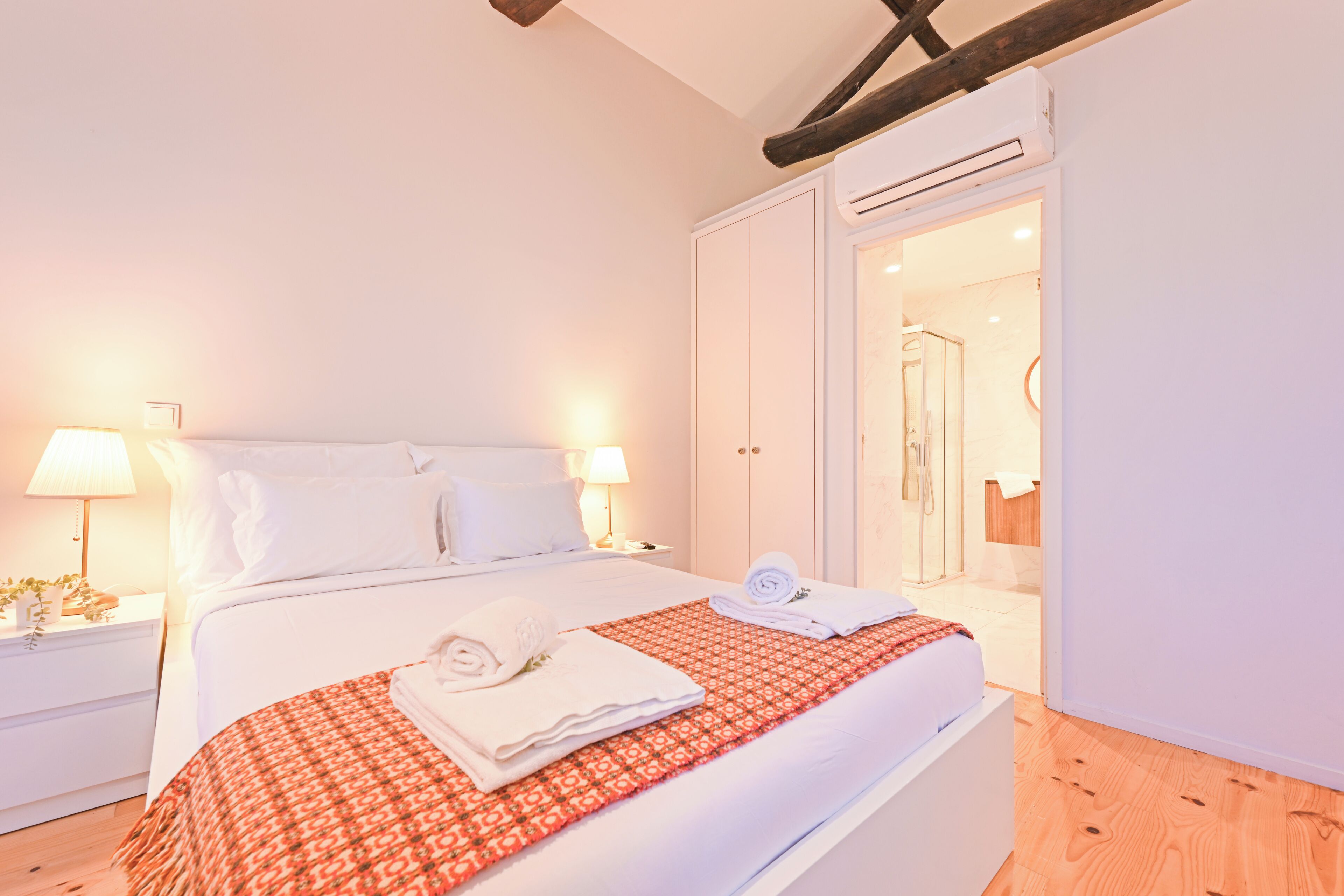 A bedroom with a bed, two bedside tables with lamps, a wardrobe, and an air conditioning unit.