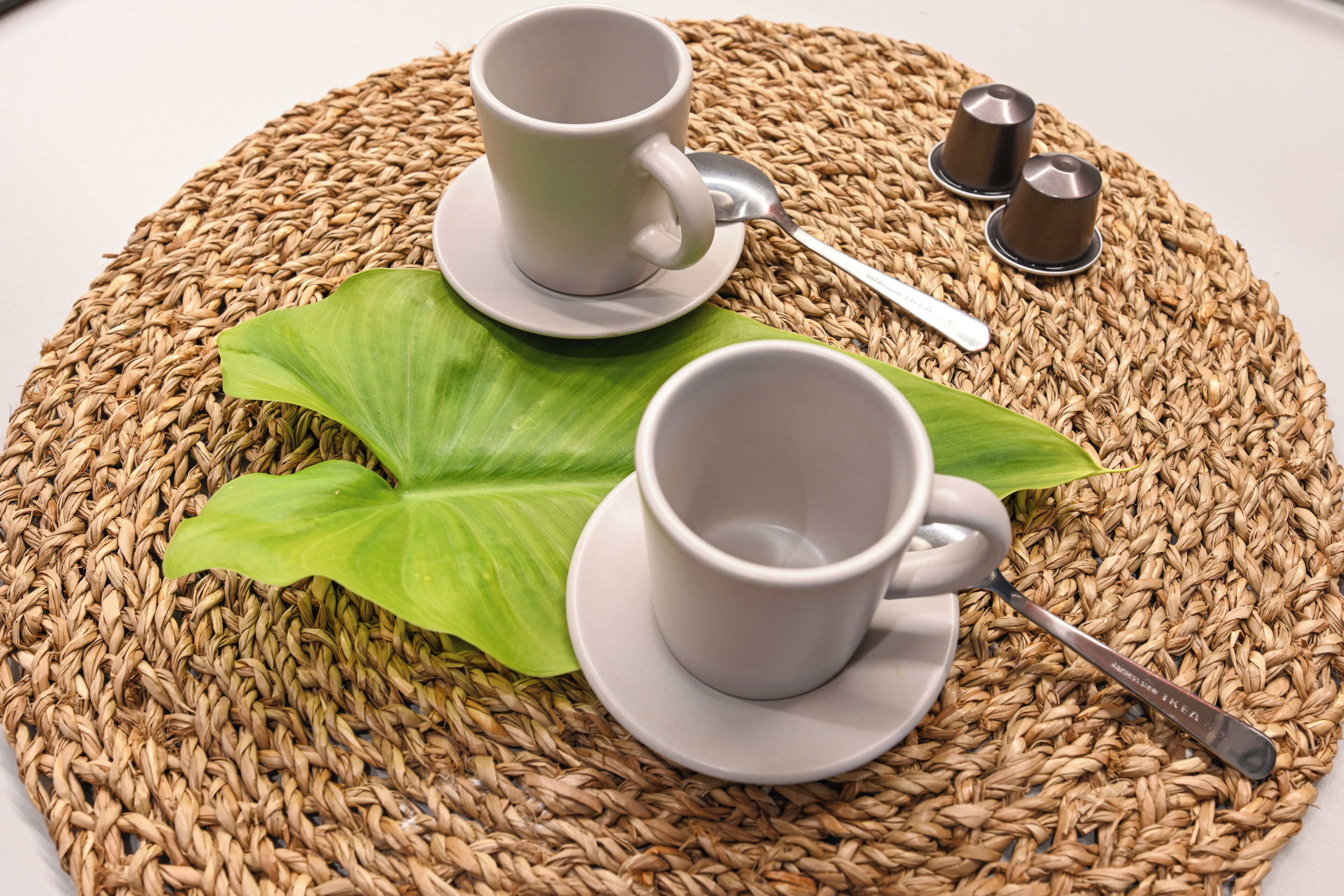 Two white cups on a woven placemat with a green leaf and a spoon.