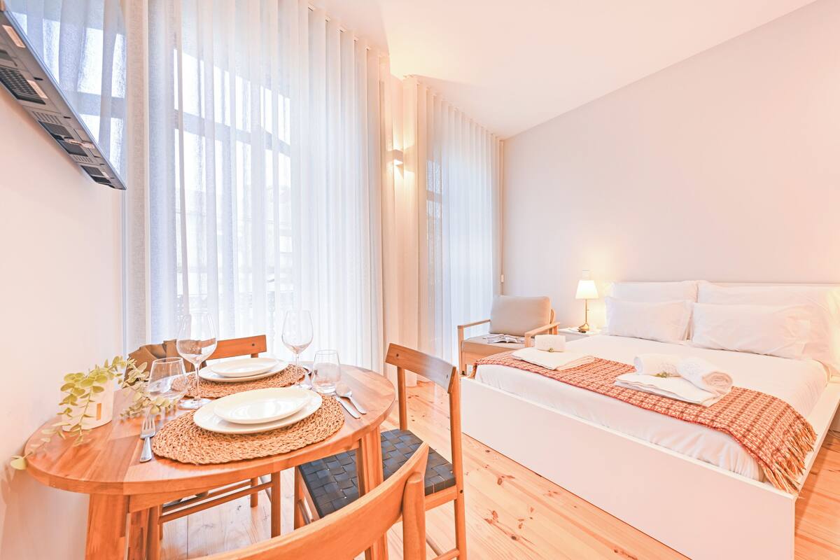 A hotel room with a bed, bedside table, lamp, and a dining area with a table set for two.