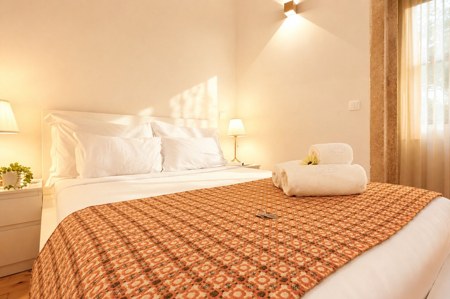 Ein Hotelzimmer mit einem Bett, einem Nachttisch mit Lampe, einem aufgerollten Handtuch und einem Fenster mit Vorhängen.