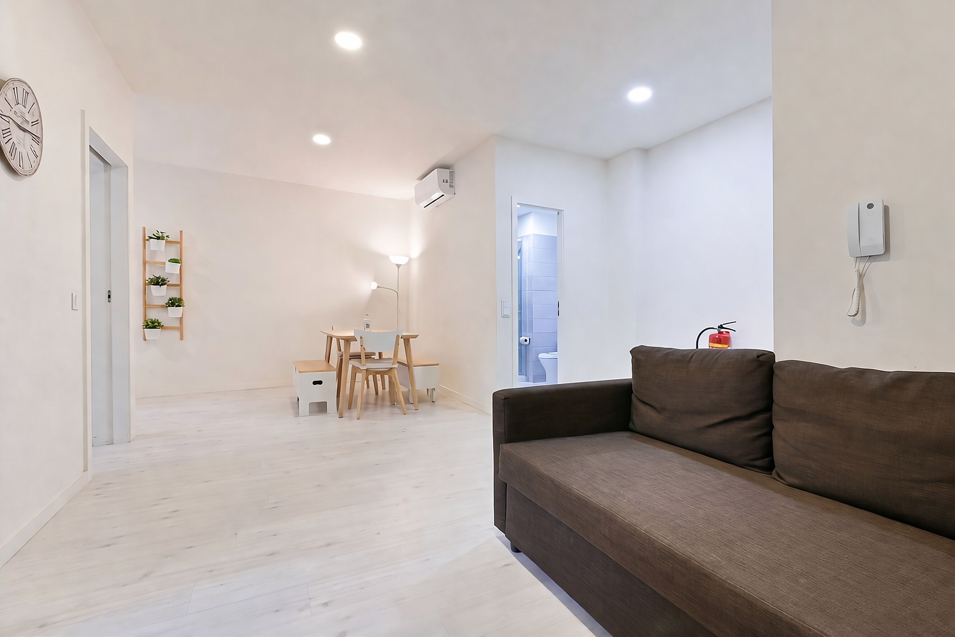 A modern living room with a brown sofa, a dining area with a table and chairs, and a hallway leading to a bathroom.