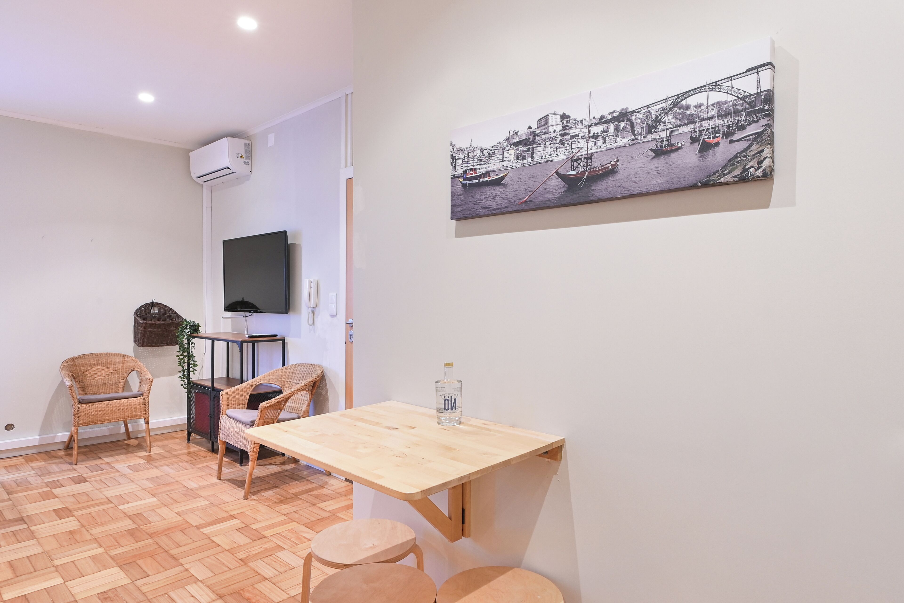 A small, cozy room with a wooden table and two chairs, a television on the wall, and a painting of a harbor scene.