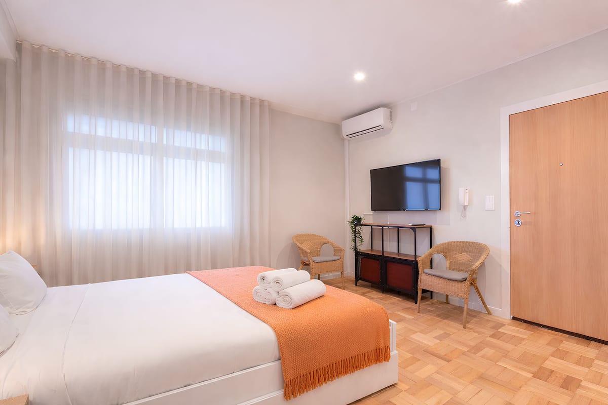 Une chambre d’hôtel avec un lit, une télévision, deux fauteuils en osier, une porte en bois et une grande fenêtre avec des rideaux.