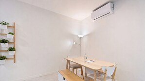Una habitación minimalista con una mesa de madera, dos sillas, un banco, estante con plantas, lámpara de escritorio y unidad de aire acondicionado.
