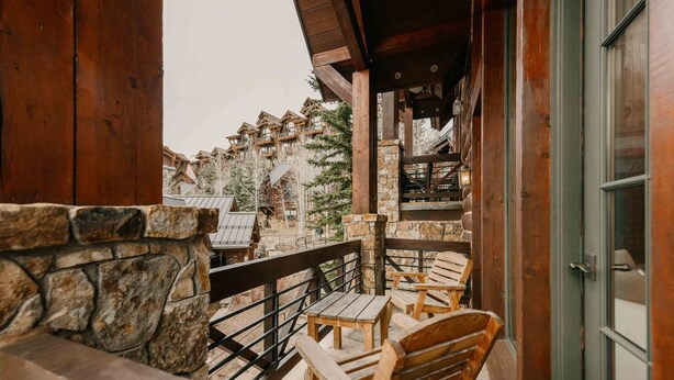 A wooden balcony with stone walls, wooden chairs, and a view of a mountain lodge with multiple buildings and trees.