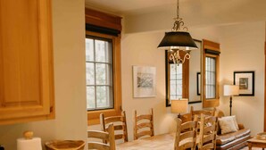A cozy kitchen with wooden cabinets, a dining table with chairs, a hanging light fixture, and a sofa in the background.