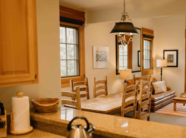 A cozy kitchen with wooden cabinets, a dining table with chairs, a hanging light fixture, and a sofa in the background. - Private See Forever Cabin by InvitedHome | Deck, Ski Valet, Walk to Ski (Mountain Village)
