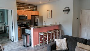 Una cocina moderna con armarios de madera, un refrigerador de acero inoxidable y un microondas. Hay una pequeña isla con taburetes y un reloj en la pared.
