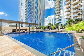A swimming pool with a wooden deck, surrounded by lounge chairs and palm trees, with tall buildings in the background.