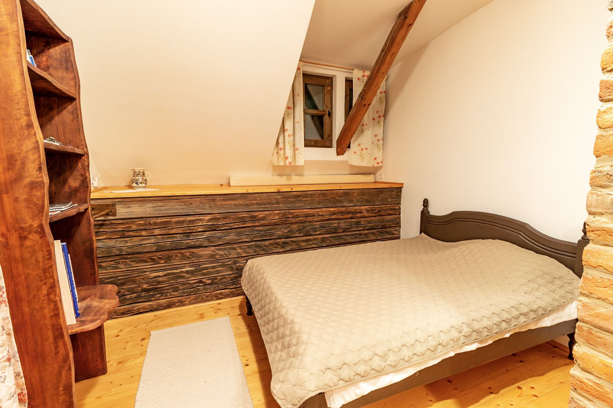 A bedroom with a wooden headboard, a bed with a quilted cover, a wooden shelf with books, and a small window with curtains.