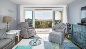 Un soggiorno con due poltrone grigie, un tavolino bianco con una lampada, un tavolino da caffè blu e un mobile contenitore color acquamarina. Un'ampia porta scorrevole in vetro dà accesso a un balcone con vista sulla spiaggia.