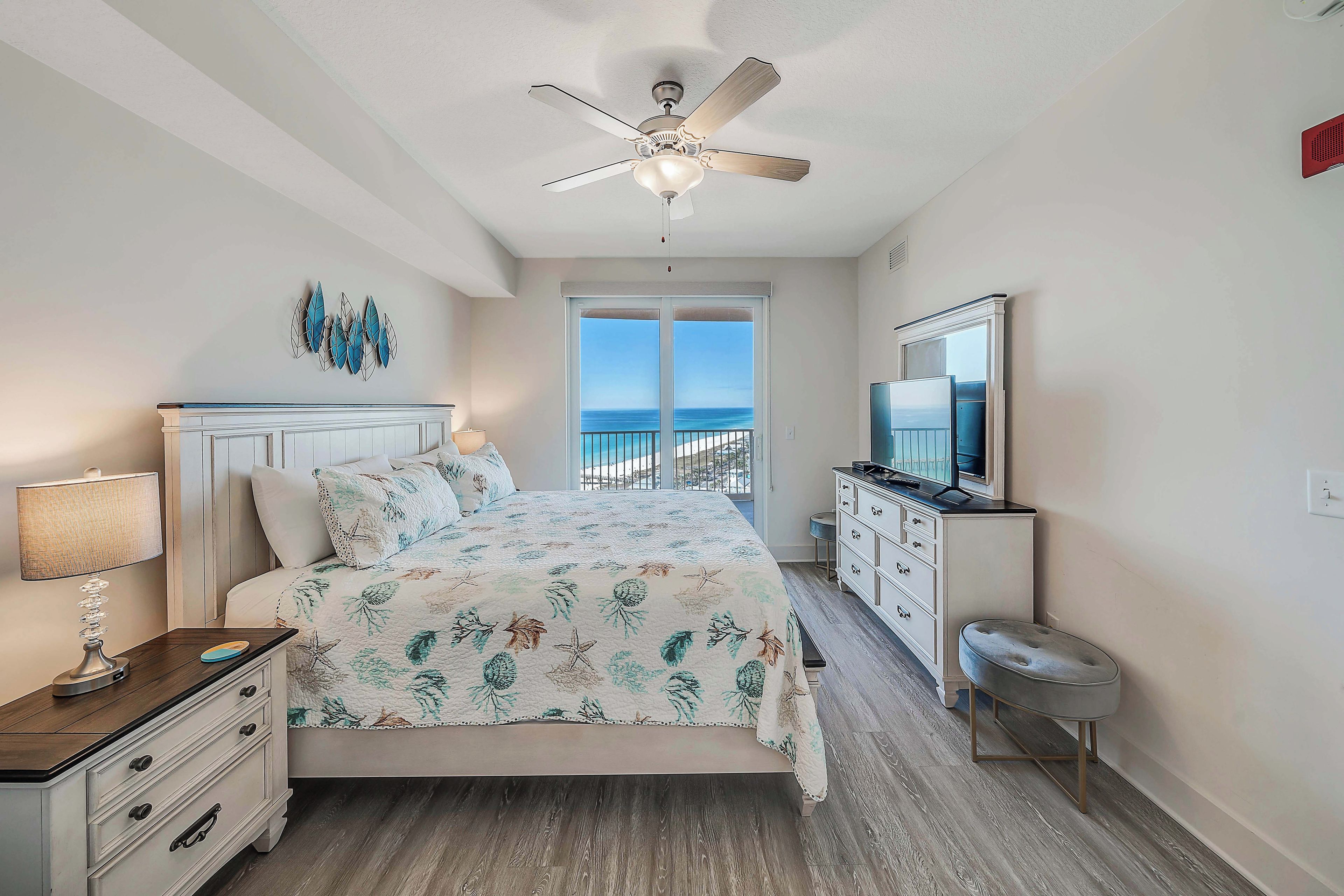 A bedroom with a bed, a ceiling fan, a dresser, a TV, and a view of the ocean through a sliding door.