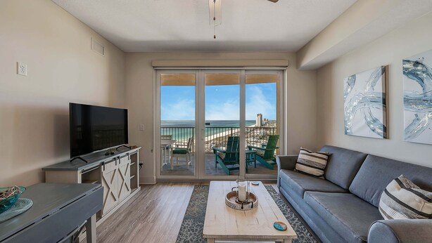 A living room with a TV, sofa, coffee table, and a view of the beach through the sliding doors. - Calypso 3-1809 Gulf View King Master, Free Golf&Perks, Pier Park, Heated Pool (Panama City Beach)