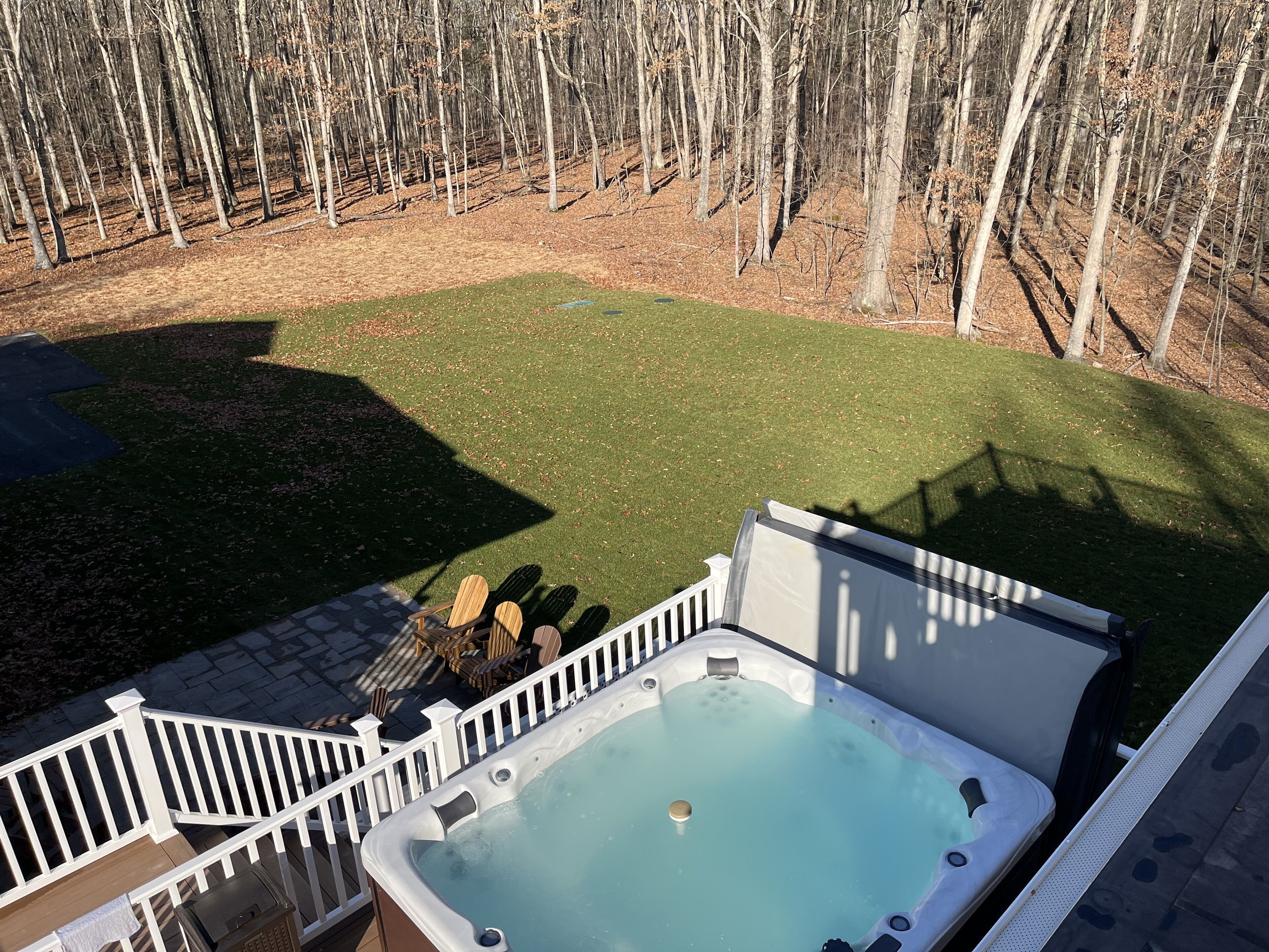 A hot tub with a wooden deck and a white railing, surrounded by a grassy yard and a wooded area.