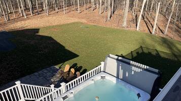 A hot tub with a wooden deck and a white railing, surrounded by a grassy yard and a wooded area.