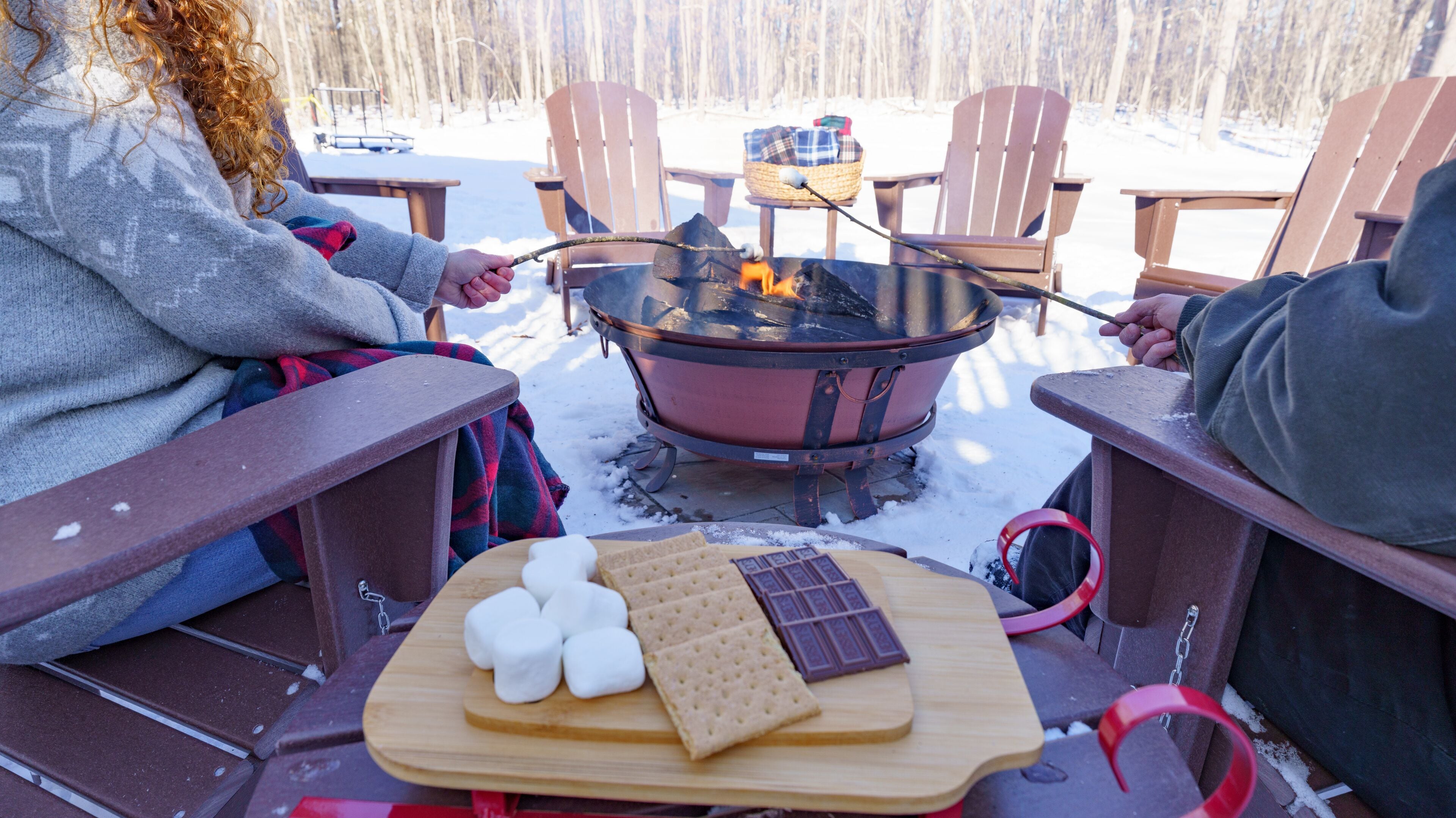 A group of people sitting on Adirondack chairs around a wood-burning stove, with marshmallows and chocolate on a board in front of them.