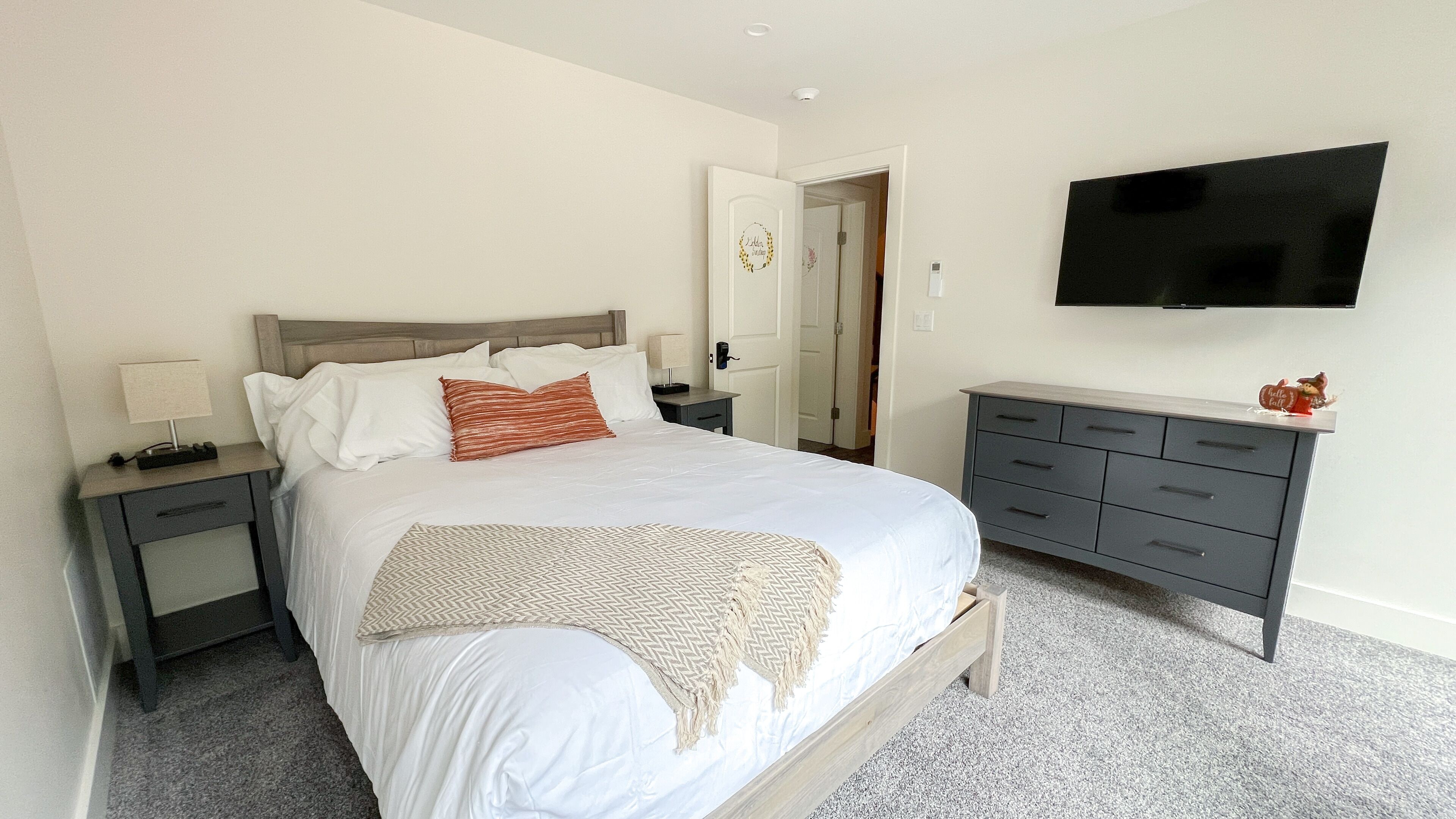 Un dormitorio ordenado con cama, mesitas de noche, cómoda y televisor de pantalla plana montado en la pared.