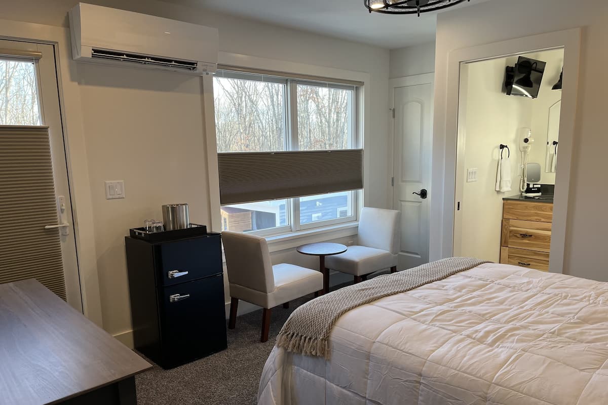 Une chambre d’hôtel comprenant un lit, un bureau, une chaise, une petite table, une armoire et une fenêtre avec des stores.