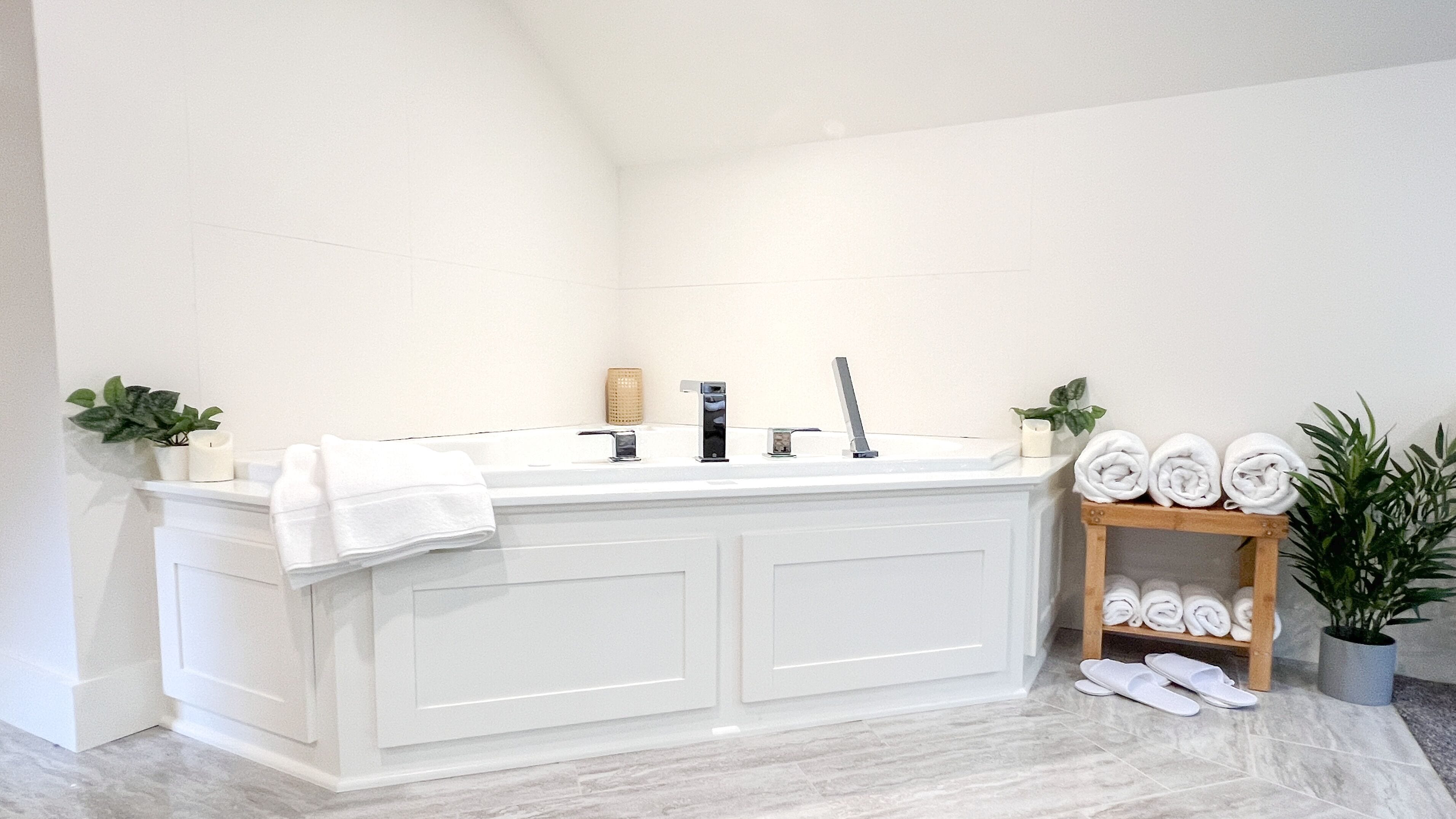 Un baño moderno con un amplio tocador blanco, una bañera independiente y un estante de madera con toallas y una planta en maceta.