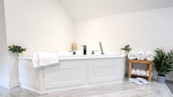 Un baño moderno con un amplio tocador blanco, una bañera independiente y un estante de madera con toallas y una planta en maceta.