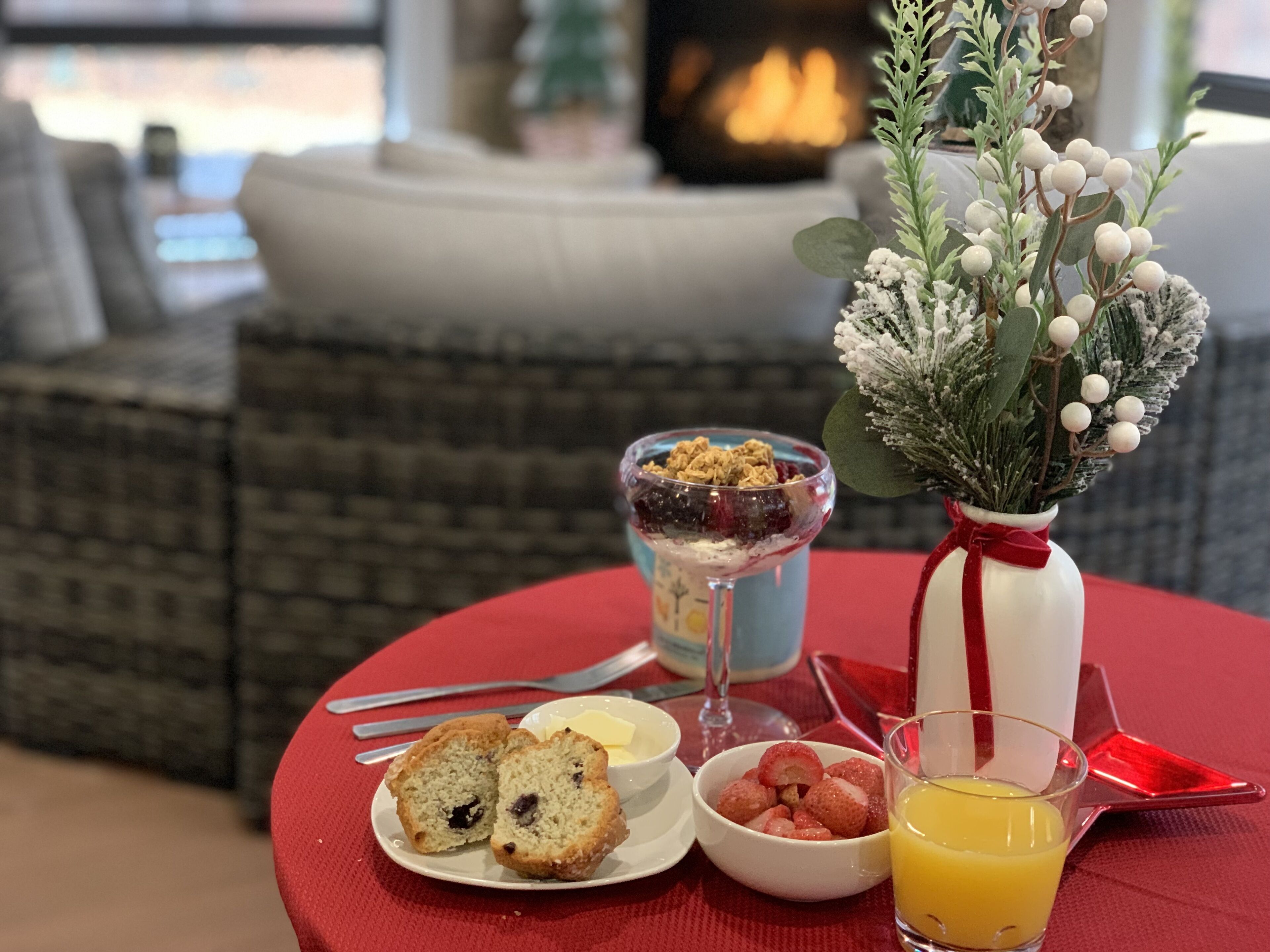 A breakfast setting with a plate of muffins, a bowl of strawberries, a glass of orange juice, and a glass of yogurt parfait on a red tablecloth.