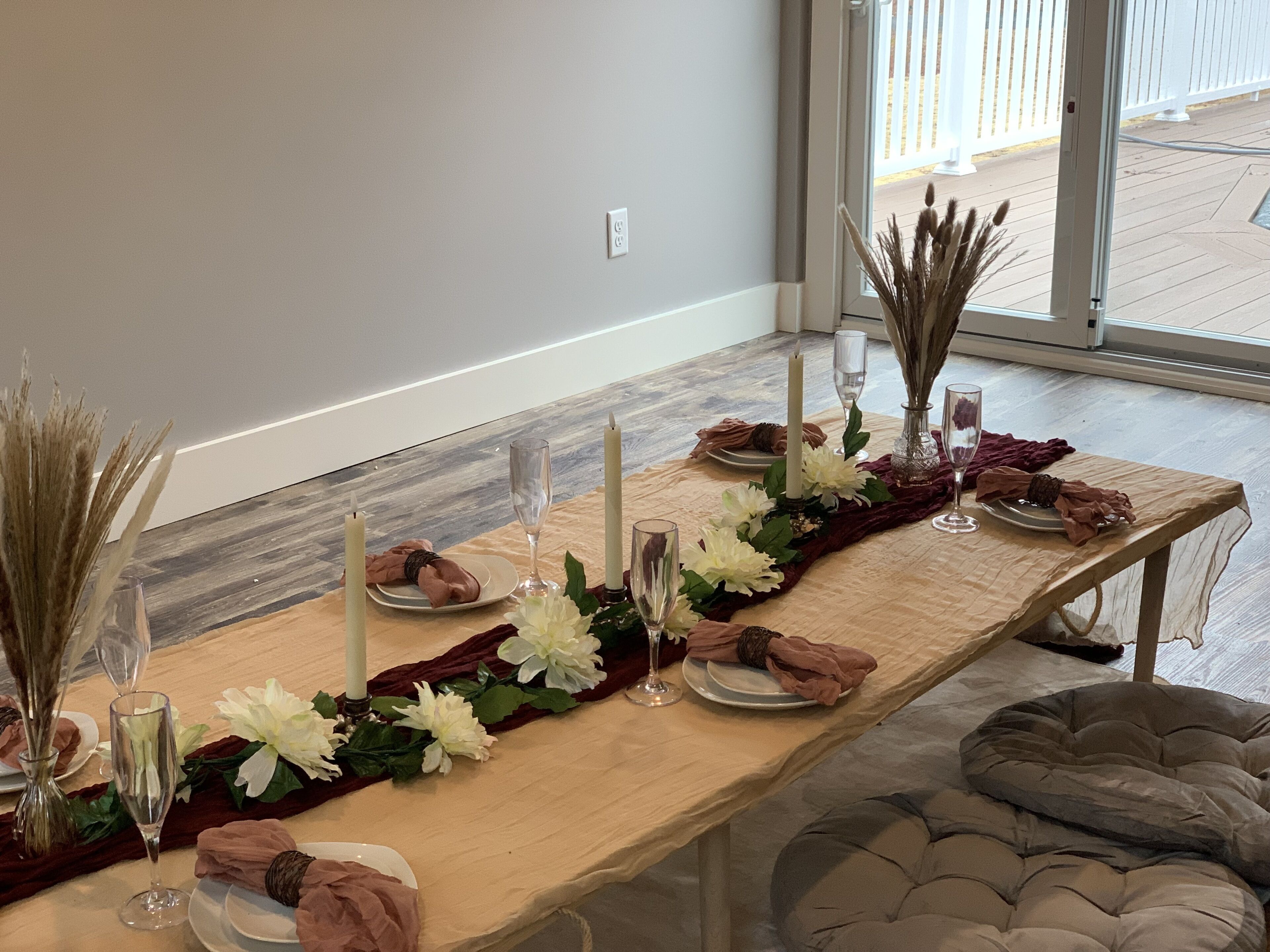 Mesa de comedor decorada con flores blancas, velas y cristalería, con vista a una terraza de madera.