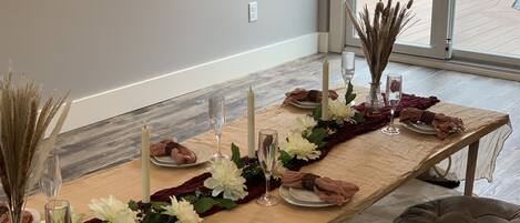 A dining table set with white flowers, candles, and glassware, with a view of a wooden deck outside.